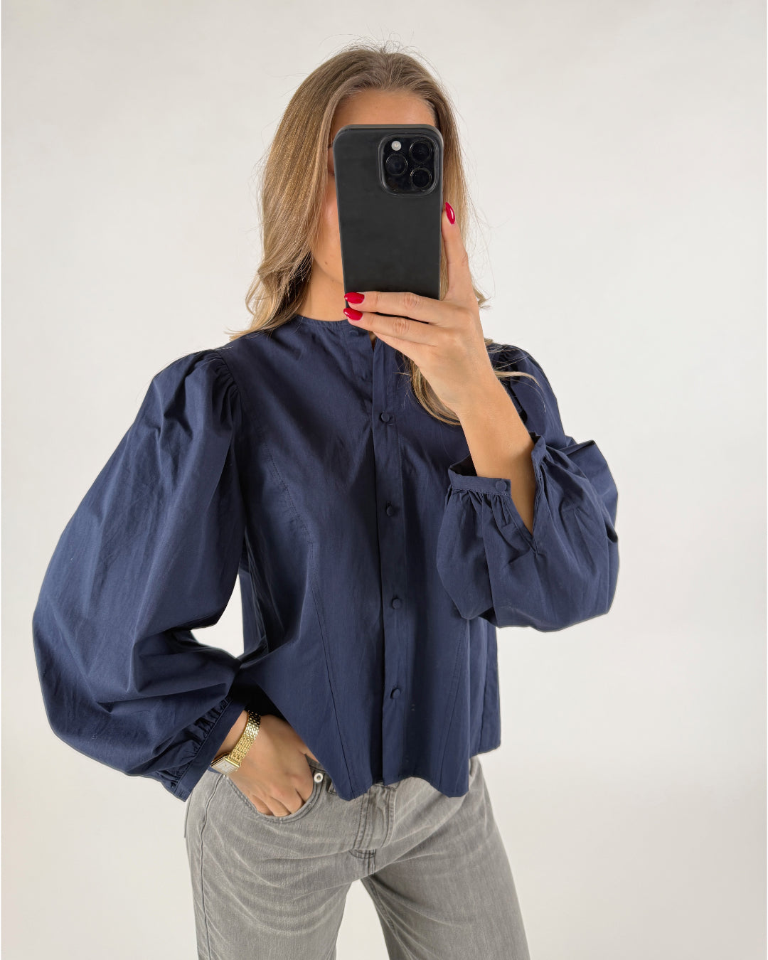 Person iført en blå bluse med pufærmer tager et spejlbillede med en sort telefon. De står i lyse jeans i en lys baggrund.