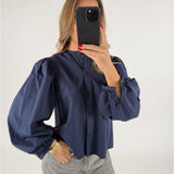 Person iført en blå bluse med pufærmer tager et spejlbillede med en sort telefon. De står i lyse jeans i en lys baggrund.