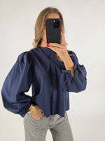 Person iført en blå bluse med pufærmer tager et spejlbillede med en sort telefon. De står i lyse jeans i en lys baggrund.