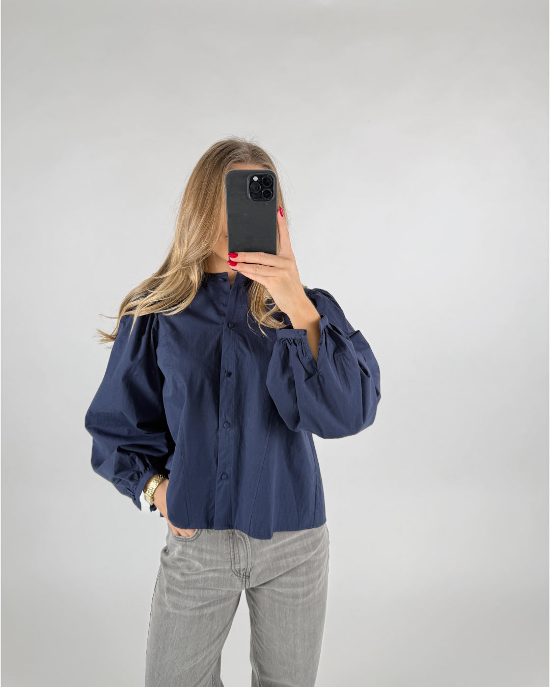 En person med langt hår tager et spejlbillede med en smartphone. De bærer en mørkeblå skjorte med pufærmer og grå jeans, stående i et neutralt rum.