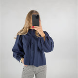 En person med langt hår tager et spejlbillede med en smartphone. De bærer en mørkeblå skjorte med pufærmer og grå jeans, stående i et neutralt rum.