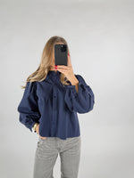 En person med langt hår tager et spejlbillede med en smartphone. De bærer en mørkeblå skjorte med pufærmer og grå jeans, stående i et neutralt rum.