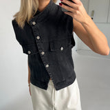 En person tager et spejlbillede iført en sort denimvest og hvide bukser med bindebånd, omgivet af en minimalistisk, lys baggrund.