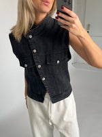 En person tager et spejlbillede iført en sort denimvest og hvide bukser med bindebånd, omgivet af en minimalistisk, lys baggrund.