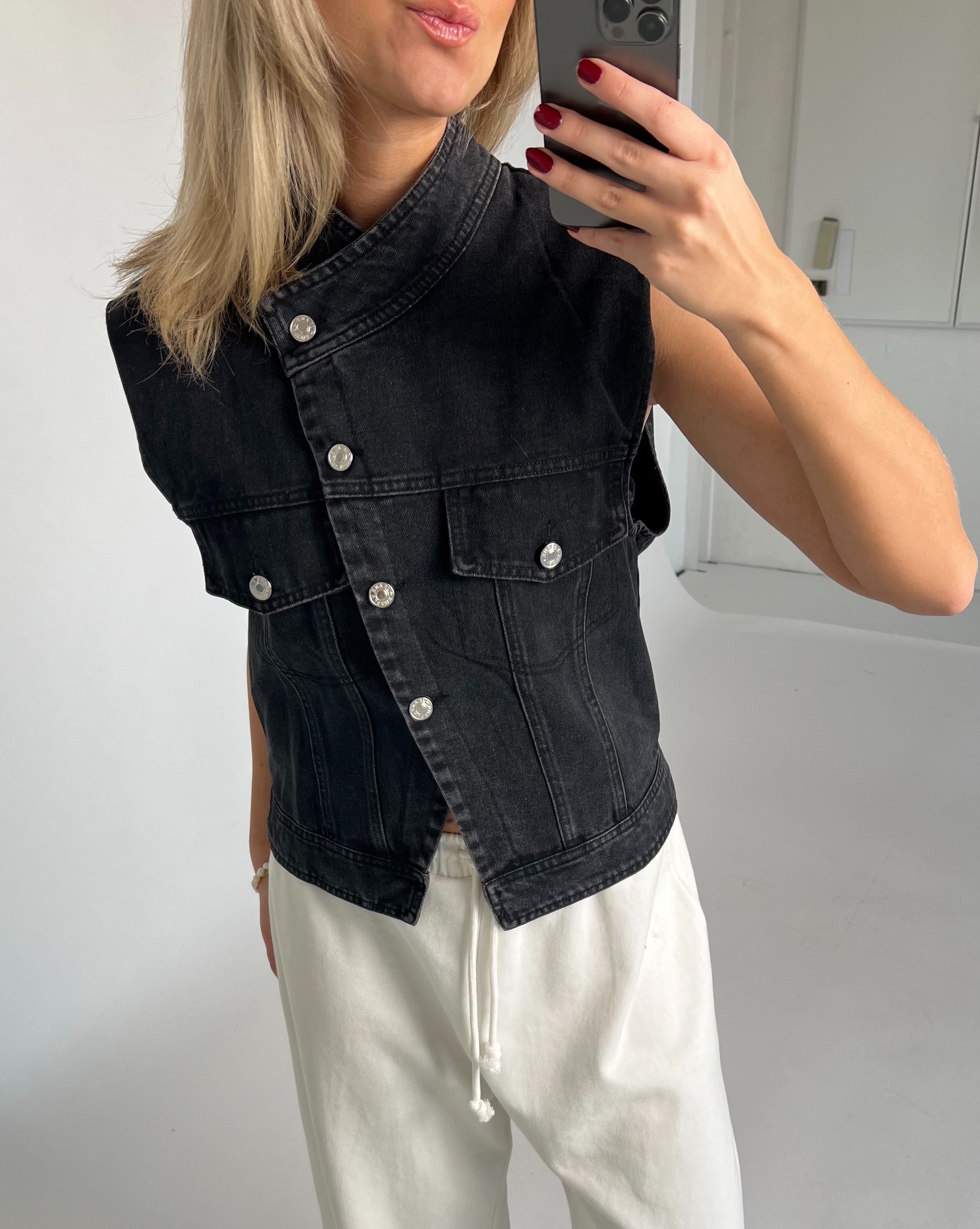 En person tager et spejlbillede iført en sort denimvest og hvide bukser med bindebånd, omgivet af en minimalistisk, lys baggrund.