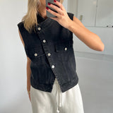 Person tager et spejlbillede iført en sort denimvest og hvide bukser. Personen holder en mobiltelefon med rødmalede negle i et rum med hvide vægge og gulv.
