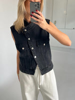 Person tager et spejlbillede iført en sort denimvest og hvide bukser. Personen holder en mobiltelefon med rødmalede negle i et rum med hvide vægge og gulv.