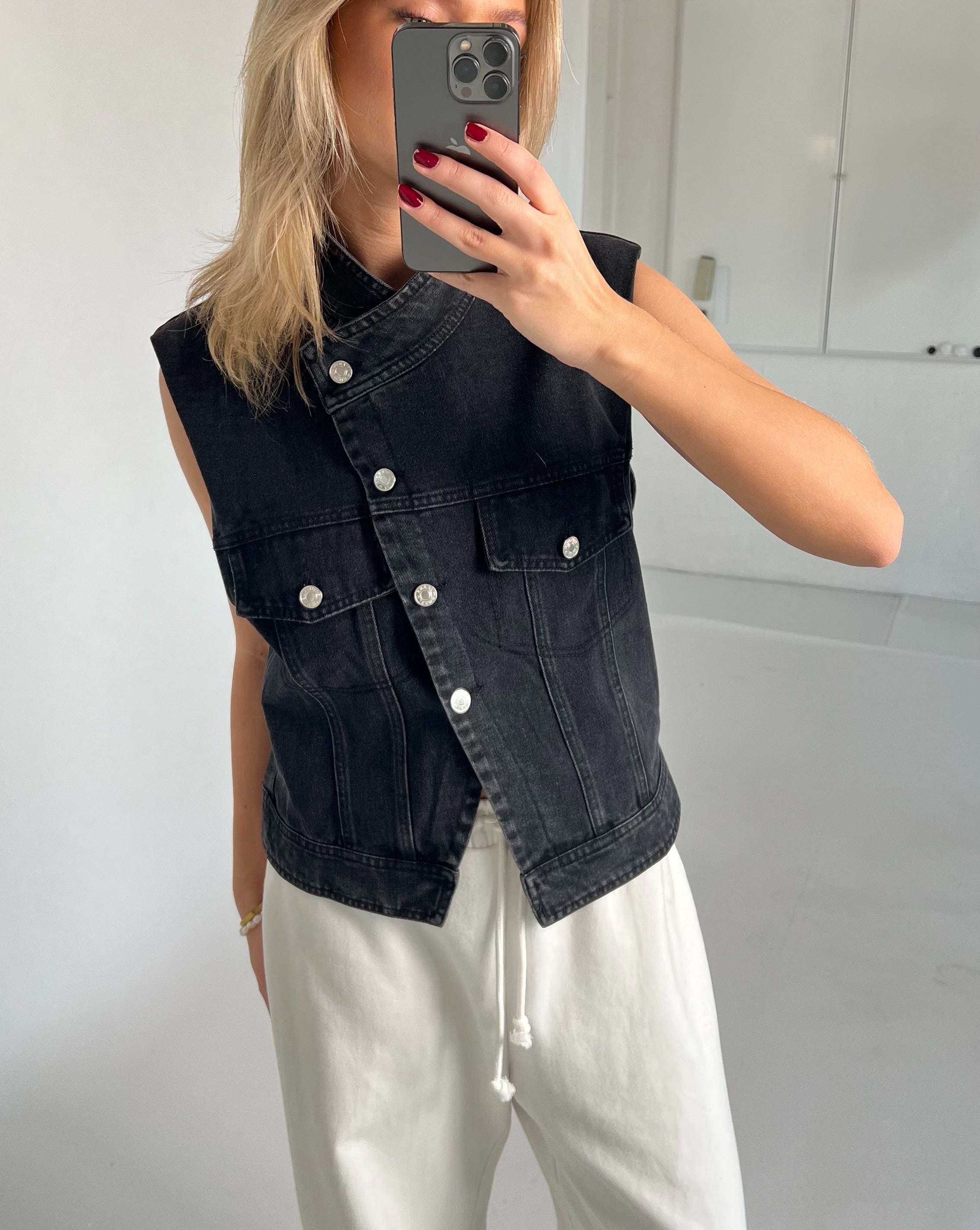 Person tager et spejlbillede iført en sort denimvest og hvide bukser. Personen holder en mobiltelefon med rødmalede negle i et rum med hvide vægge og gulv.