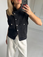 En person iført en sort denimvest med knapper tager et spejlfoto. Personen har hvide bukser på og står i et lyst rum.