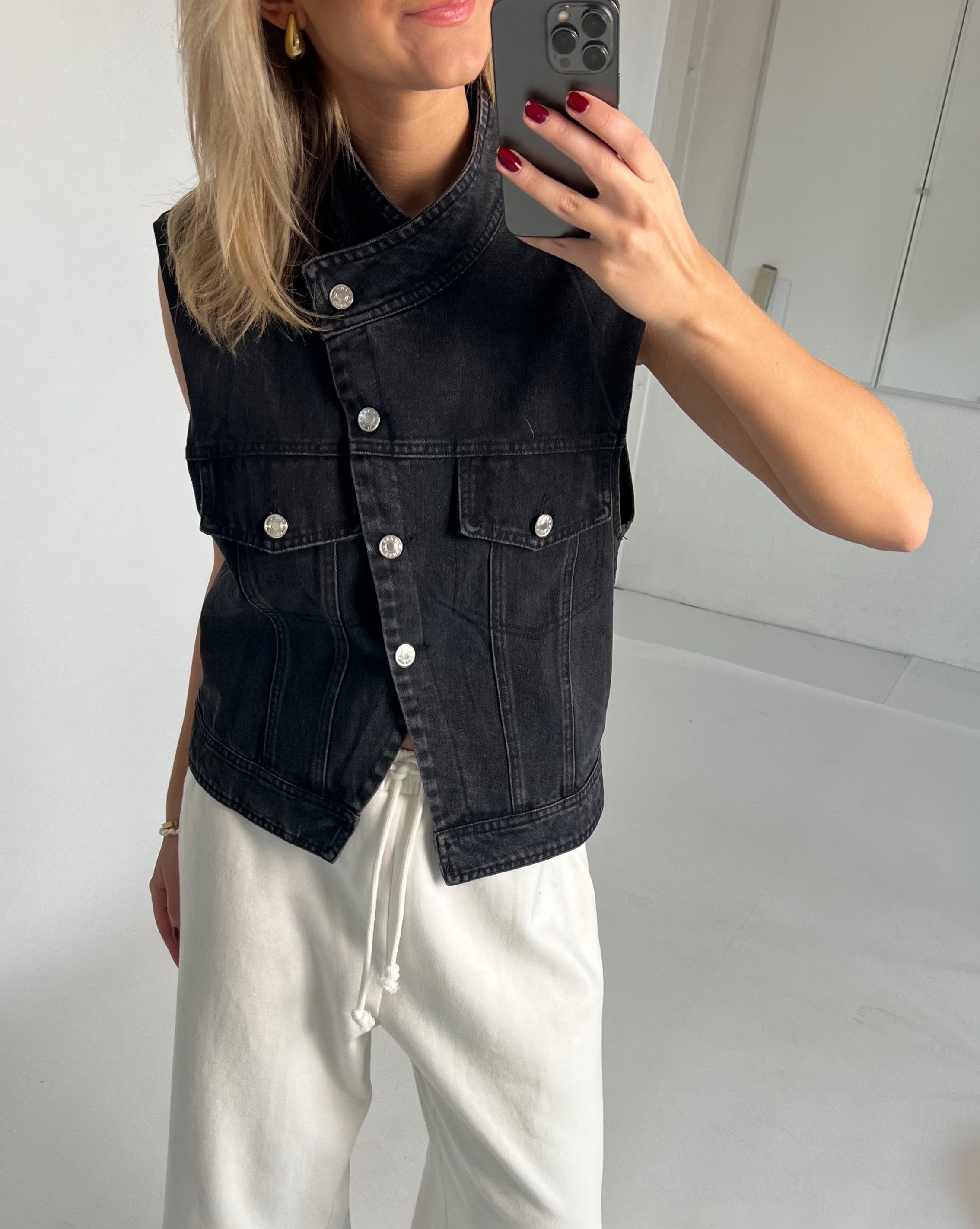 En person iført en sort denimvest med knapper tager et spejlfoto. Personen har hvide bukser på og står i et lyst rum.