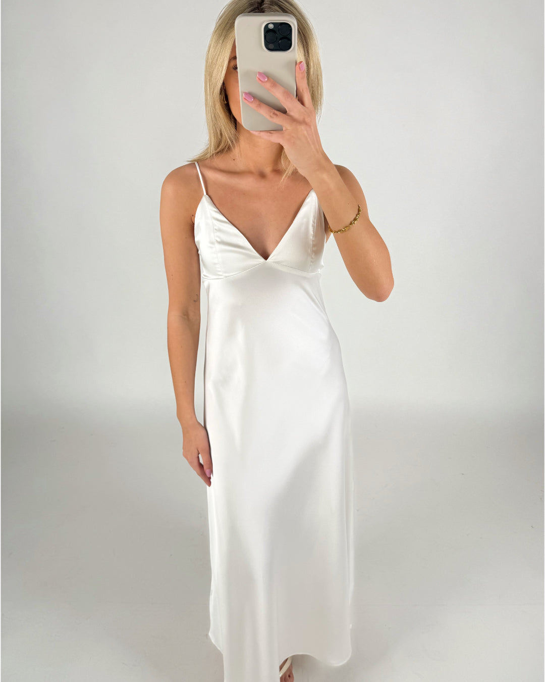 En kvinde iført en hvid, lang kjole tager et spejlbillede med en mobiltelefon. Hun står i et minimalistisk, neutralt rum.