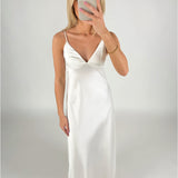 En kvinde iført en hvid, lang kjole tager et spejlbillede med en mobiltelefon. Hun står i et minimalistisk, neutralt rum.