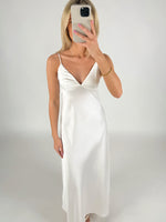 En kvinde iført en hvid, lang kjole tager et spejlbillede med en mobiltelefon. Hun står i et minimalistisk, neutralt rum.