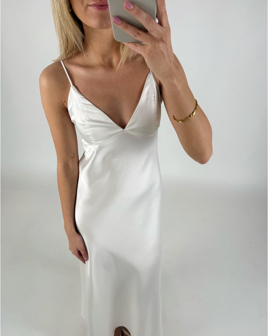 En person iført en hvid satin kjole tager et selfie med en smartphone i et lyst rum. Personen bærer et gyldent armbånd på højre håndled.