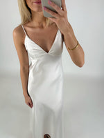 En person iført en hvid satin kjole tager et selfie med en smartphone i et lyst rum. Personen bærer et gyldent armbånd på højre håndled.