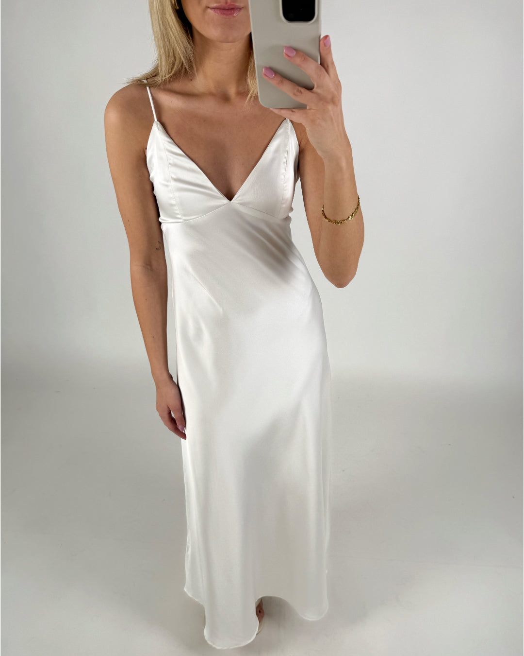 En person iført en lang, hvid kjole tager et spejlbillede. Personen holder en smartphone i et lyst rum, og kjolen har tynde stropper og en dyb udskæring.