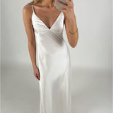 En person iført en lang, hvid kjole tager et spejlbillede. Personen holder en smartphone i et lyst rum, og kjolen har tynde stropper og en dyb udskæring.