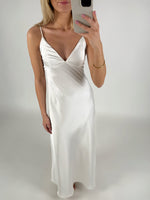 En person iført en lang, hvid kjole tager et spejlbillede. Personen holder en smartphone i et lyst rum, og kjolen har tynde stropper og en dyb udskæring.