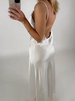 En kvinde i hvid, rygfri kjole tager et spejlbillede med en smartphone i hånden i et neutralt, hvidt rum.