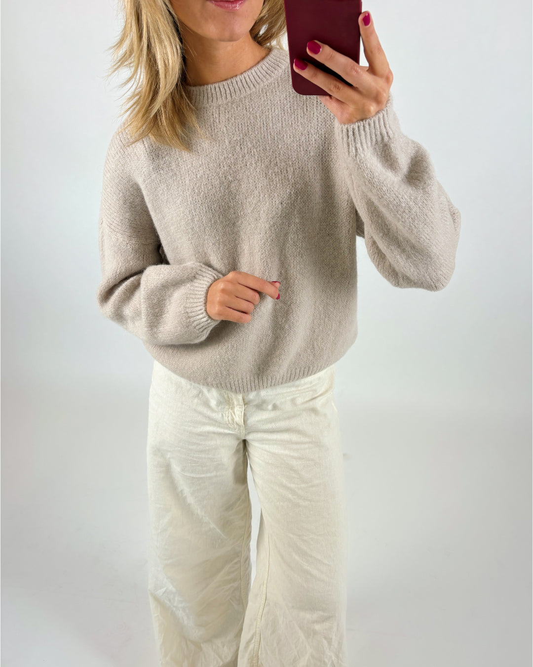En person iført en lysegrå sweater og hvide bukser tager et spejlbillede, mens de holder en rød telefon. Personen har skulderlangt, lyst hår. Baggrunden er simpel og hvid.