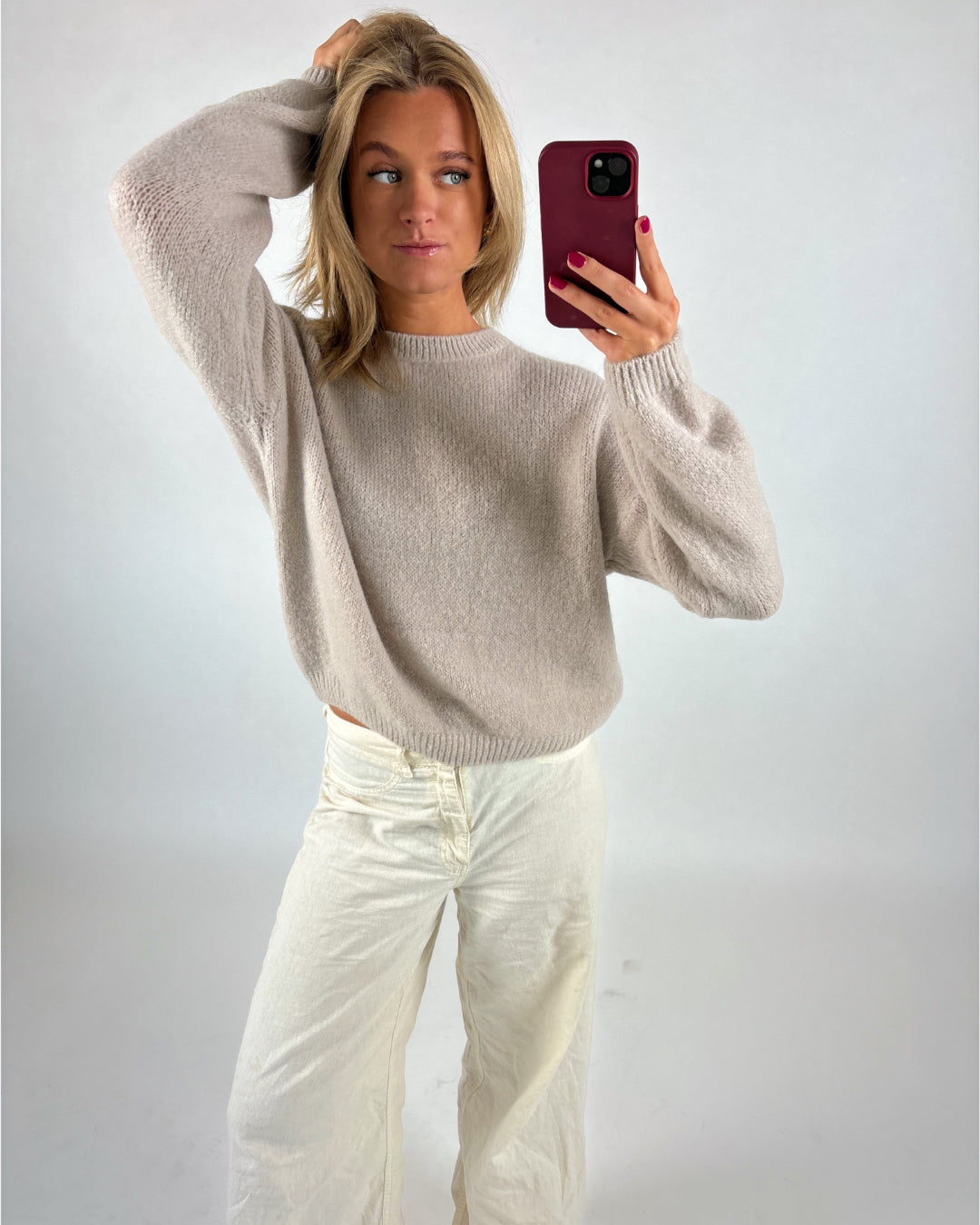 Kvinde tager selfie, iført lys beige striktrøje og hvide bukser, står i et lyst rum med én hånd i håret og den anden holder en mobiltelefon i rød etui.