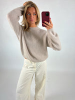 Kvinde tager selfie, iført lys beige striktrøje og hvide bukser, står i et lyst rum med én hånd i håret og den anden holder en mobiltelefon i rød etui.