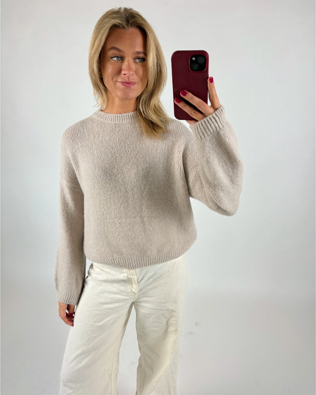 En person tager en selfie iført en lys sweater og hvide bukser, mens de står foran en ensfarvet baggrund. Personen holder en telefon med et rødt cover.