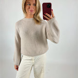 En person tager en selfie iført en lys sweater og hvide bukser, mens de står foran en ensfarvet baggrund. Personen holder en telefon med et rødt cover.