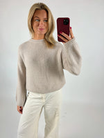 En person tager en selfie iført en lys sweater og hvide bukser, mens de står foran en ensfarvet baggrund. Personen holder en telefon med et rødt cover.