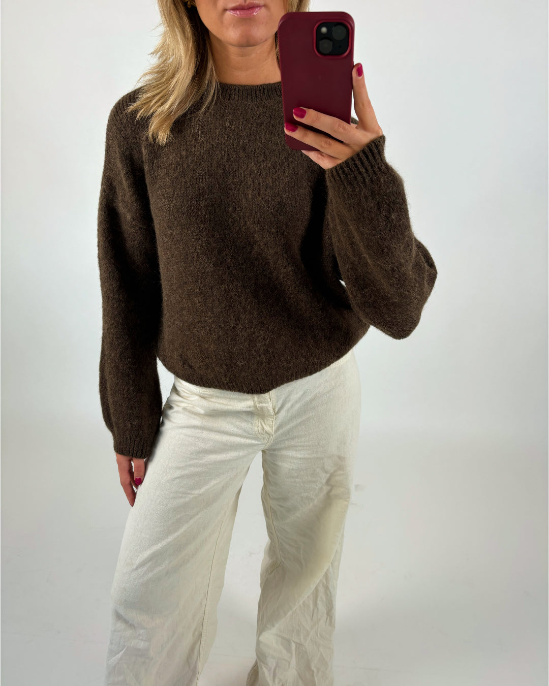En person iført en brun sweater og hvide bukser tager et spejlbillede med en mobiltelefon med rød omslag. Baggrunden er en simpel hvid væg.