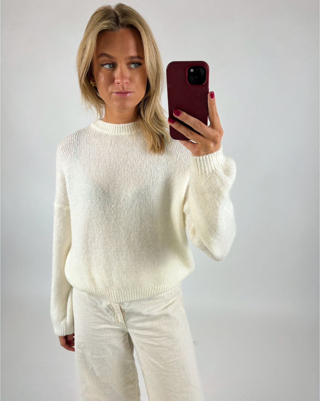 En person iført en hvid sweater og bukser tager en selfie med en mobiltelefon i en rød cover i et neutralt rum.