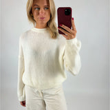 En person iført en hvid sweater og bukser tager en selfie med en mobiltelefon i en rød cover i et neutralt rum.