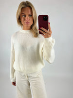En person iført en hvid sweater og bukser tager en selfie med en mobiltelefon i en rød cover i et neutralt rum.