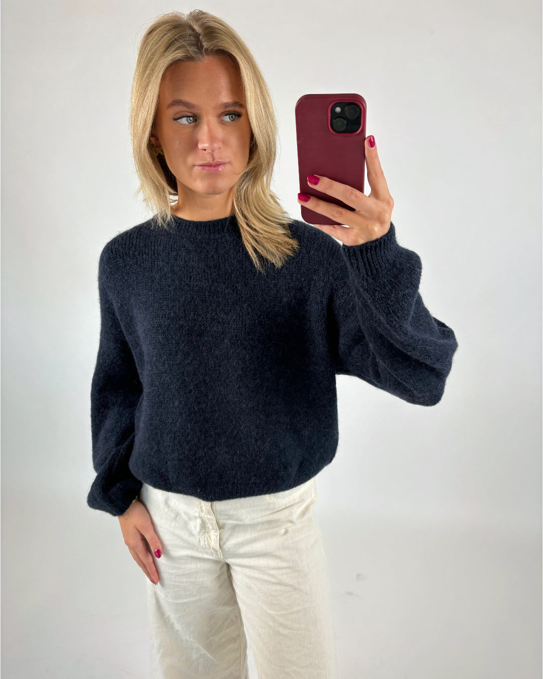 Kvinde tager et spejlbillede med mobiltelefon. Hun har en mørk sweater og lyse bukser på. Baggrunden er en simpel, lys væg.