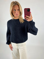 Kvinde tager et spejlbillede med mobiltelefon. Hun har en mørk sweater og lyse bukser på. Baggrunden er en simpel, lys væg.