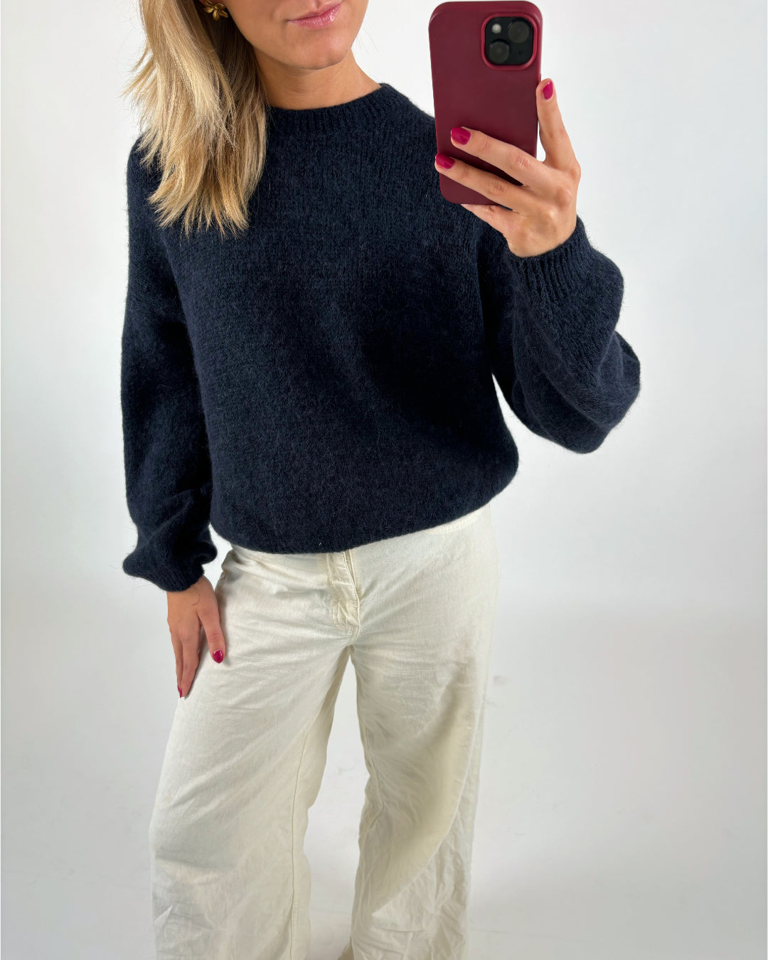 En person iført mørk sweater og lyse bukser tager et spejlbillede med en rød mobiltelefon i en minimalistisk, lys baggrund.