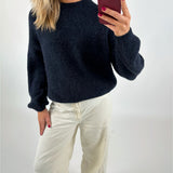 En person iført mørk sweater og lyse bukser tager et spejlbillede med en rød mobiltelefon i en minimalistisk, lys baggrund.