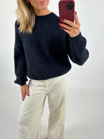 En person iført mørk sweater og lyse bukser tager et spejlbillede med en rød mobiltelefon i en minimalistisk, lys baggrund.
