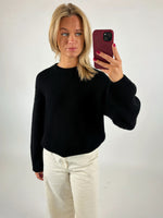 En person iført en sort trøje og hvide bukser tager en selfie med en rød telefon i et neutralt oplyst rum.