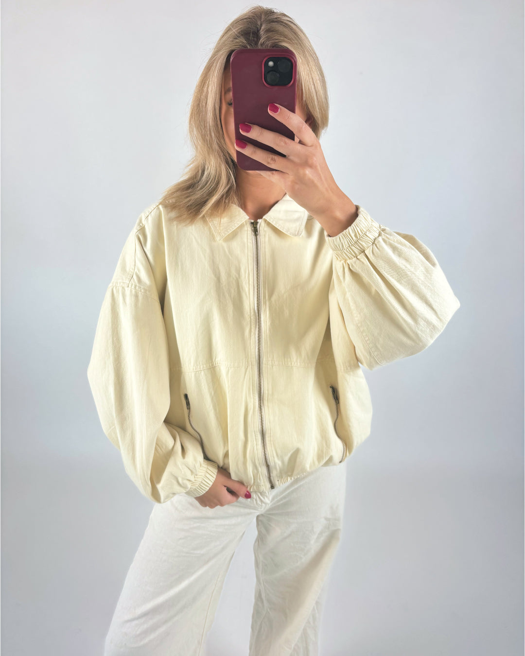 En person iført en lysegul jakke tager et selfie med en rød mobiltelefon, stående foran en hvid baggrund.