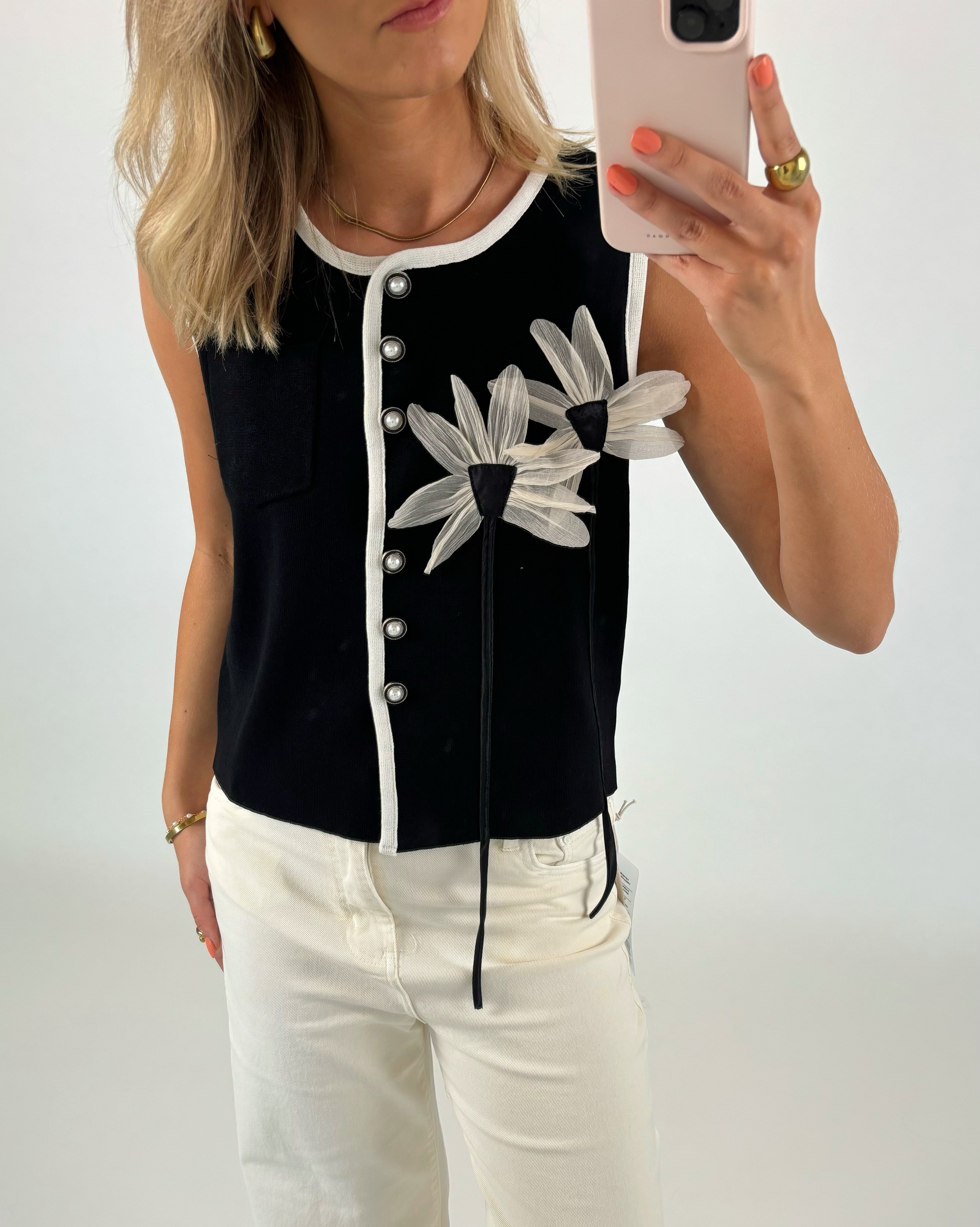 En kvinde iført en sort vest med hvide kanter og blomstermotiver tager et spejlbillede. Hun har lyst hår og bærer lyse bukser og smykker.
