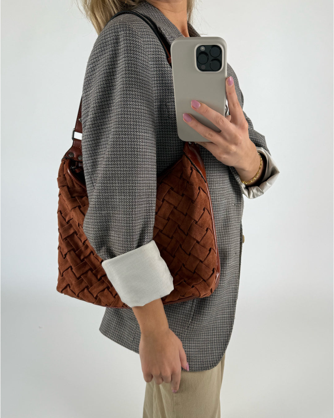 En kvinde holder en smartphone op foran sig, med en brun, vævet taske hængende over skulderen. Hun står i en grå blazer og beige bukser, mod en lys baggrund.