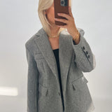 En person iført en grå jakke tager en selfie med en smartphone i en brun cover. Personen har lyst hår og står i et neutralt oplyst rum.