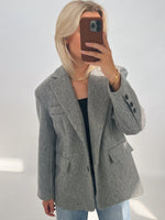 En person iført en grå jakke tager en selfie med en smartphone i en brun cover. Personen har lyst hår og står i et neutralt oplyst rum.