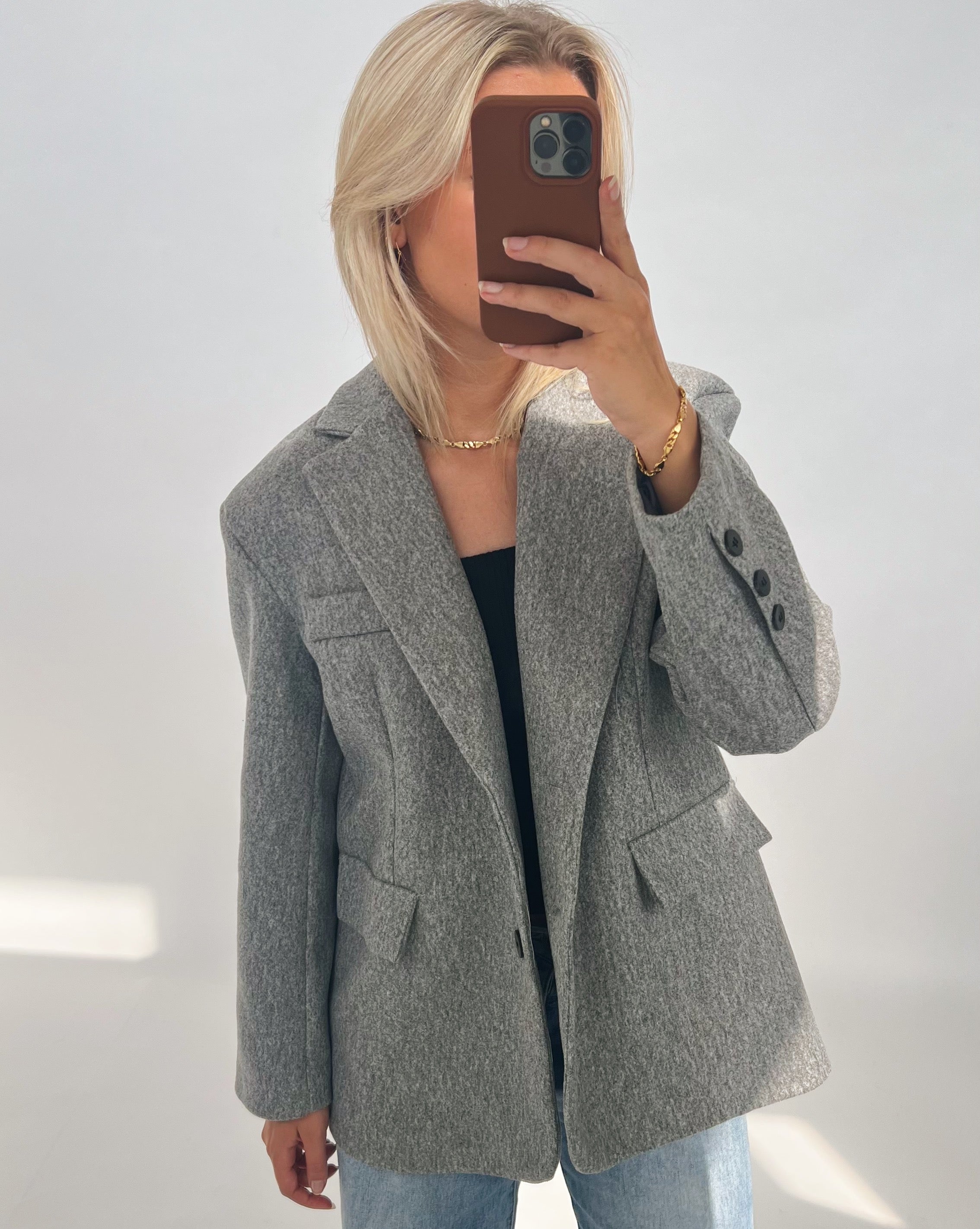 En person iført en grå jakke tager en selfie med en smartphone i en brun cover. Personen har lyst hår og står i et neutralt oplyst rum.
