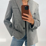 En person iført en grå blazer og blå jeans tager et spejlbillede med en smartphone i brun etui. Baggrunden er neutral med lys belysning.