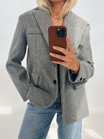 En person iført en grå blazer og blå jeans tager et spejlbillede med en smartphone i brun etui. Baggrunden er neutral med lys belysning.