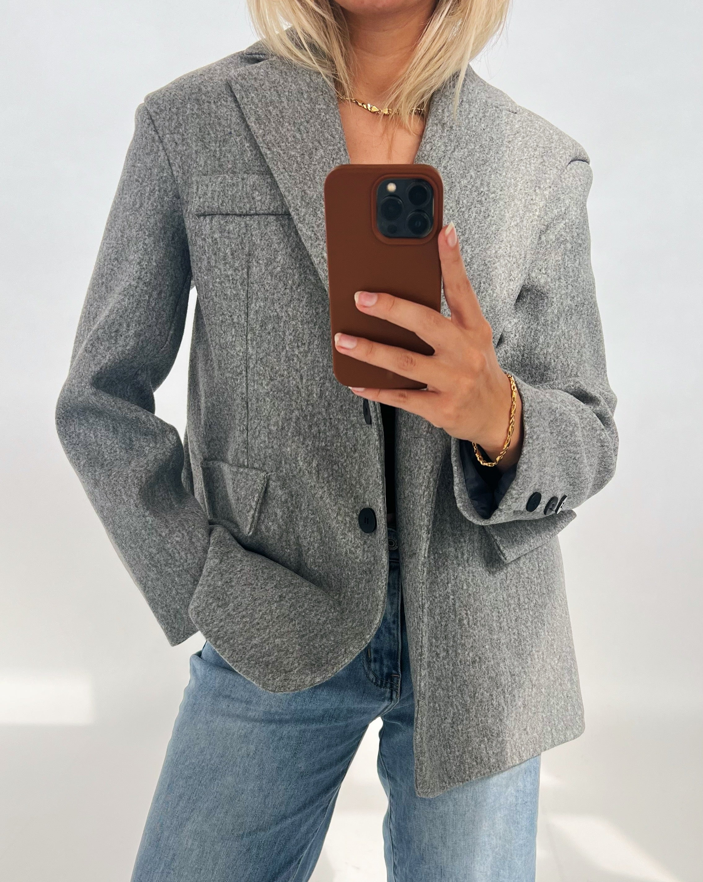 En person iført en grå blazer og blå jeans tager et spejlbillede med en smartphone i brun etui. Baggrunden er neutral med lys belysning.