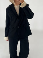 En person iført sort blazer og bukser med en lys sweater under står mod en neutral baggrund. Hånden er løftet nær ansigtet, som ikke er synligt.
