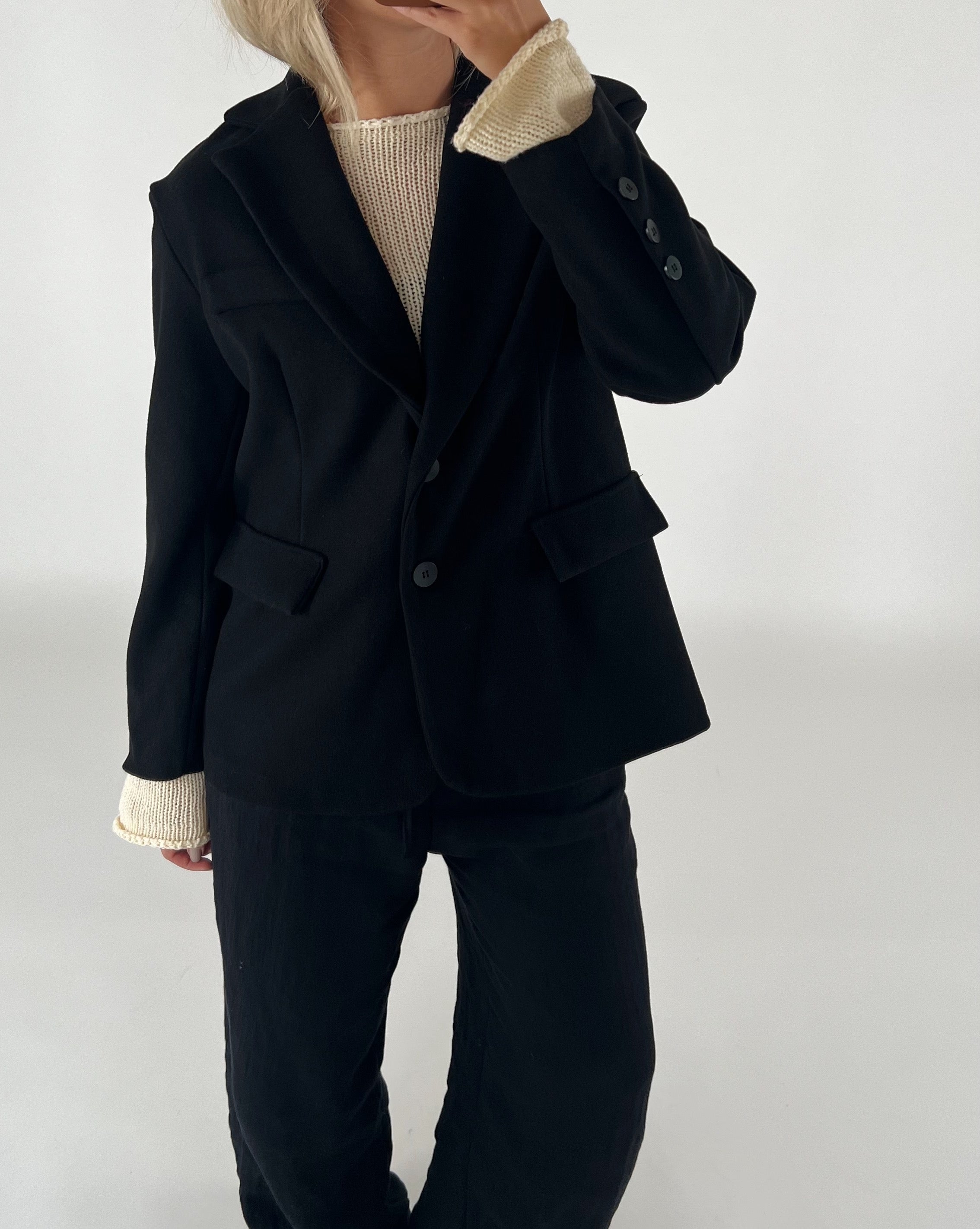 En person iført sort blazer og bukser med en lys sweater under står mod en neutral baggrund. Hånden er løftet nær ansigtet, som ikke er synligt.